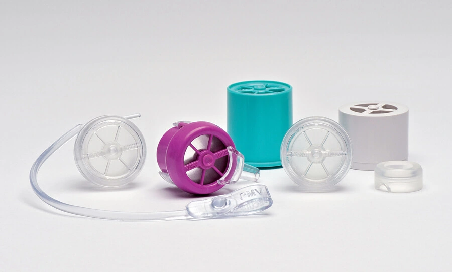 A collection of tracheostomy speaking valves in various colors and designs, including a purple valve, teal valve, and clear transparent valves, arranged on a neutral background